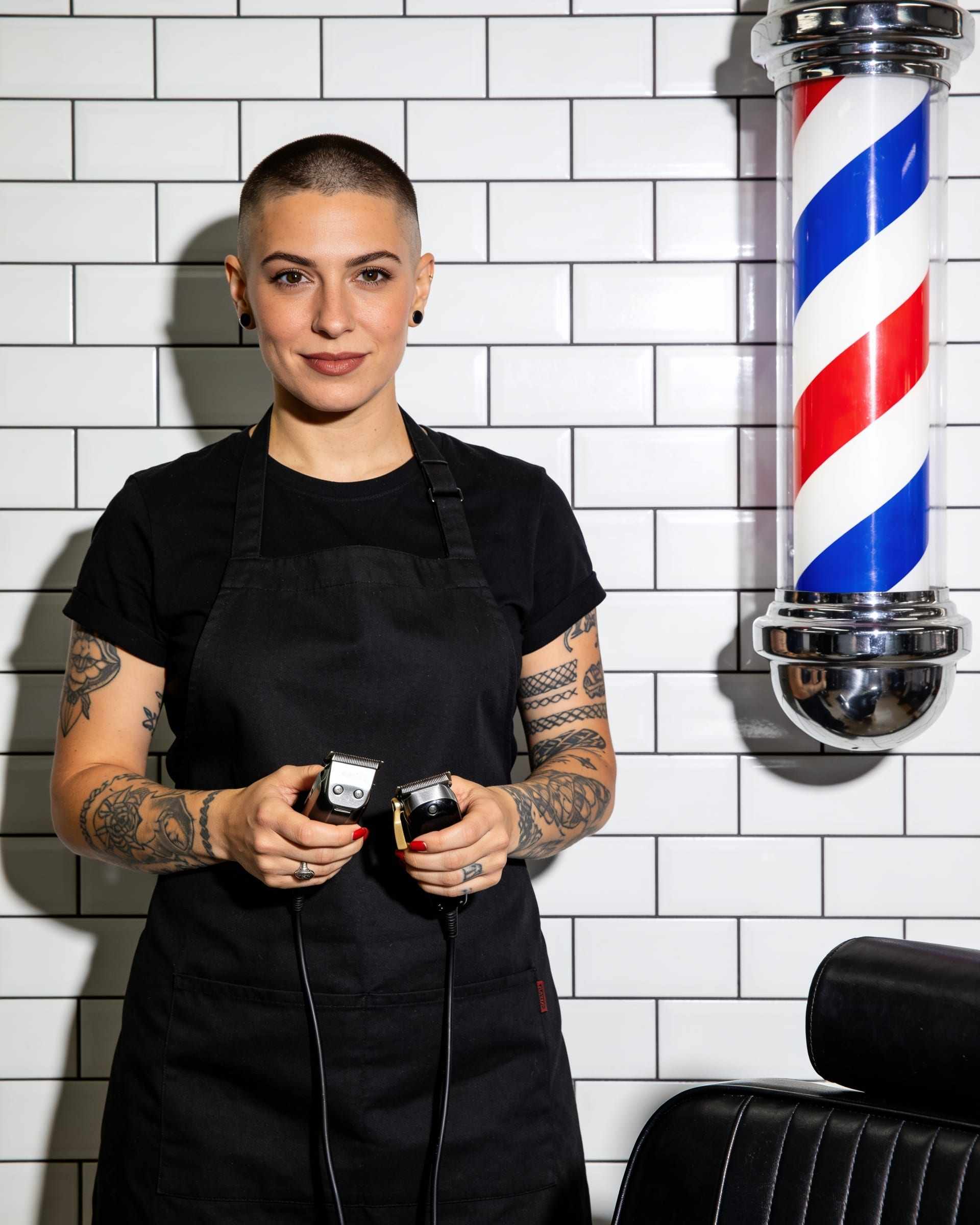 A barber with a short undercut and tattooed forearms in a black apron, holding clippers, against white tiles